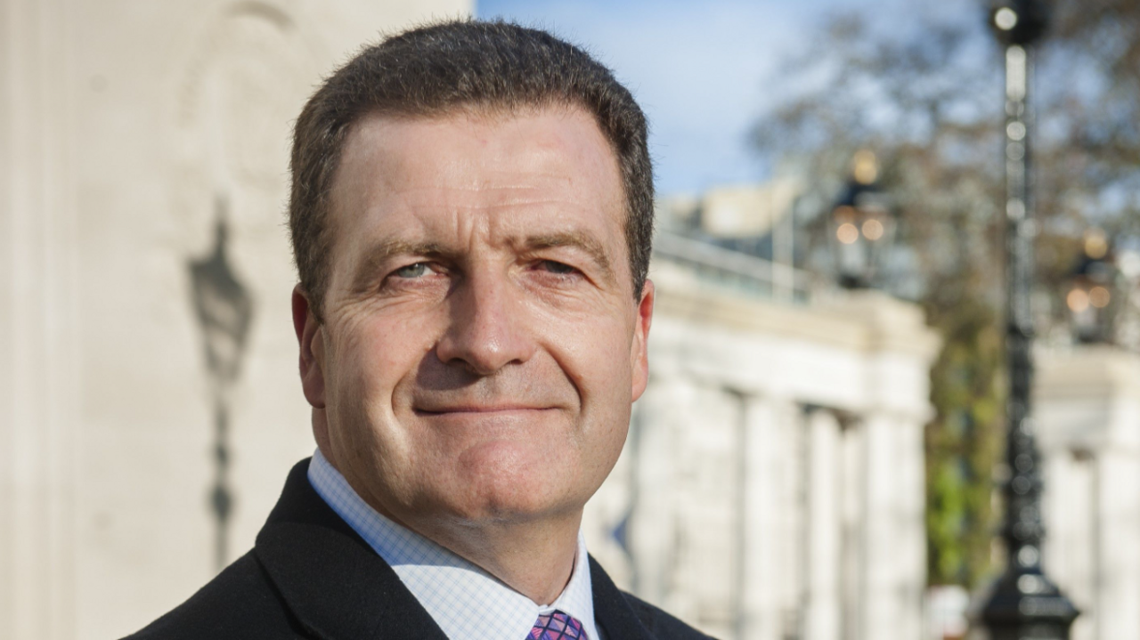 John Nichol wearing a black jacket, white and blue chequered shirt and tie