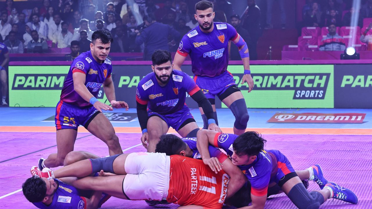 Men playing kabaddi and dressed in purple, white and red. One in a red T-shirt and white shorts, is lying on the ground, held by three other players in purple and white, while three of their team-mates stand behind them, looking on