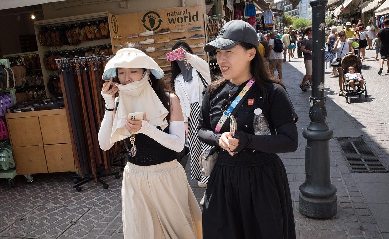 Two people wearing sun hats and sun coverings walk down a street carrying water, with people in the background, in Crete on 21 August