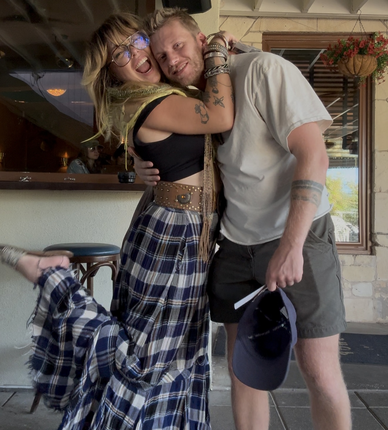 Jessica, wearing a long blue checked skirt and black sleeveless top, hugs her partner who is wearing dark grey shorts and a light t-shirt and holding a blue cap.