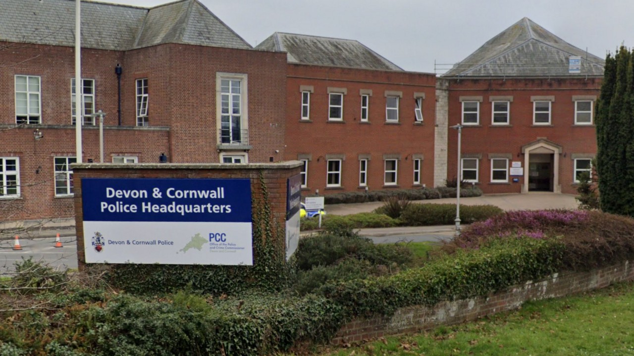 Devon and Cornwall Police Headquarters is written on a sign that stands in front of a two-storey red brick building with shrubs in the foreground.