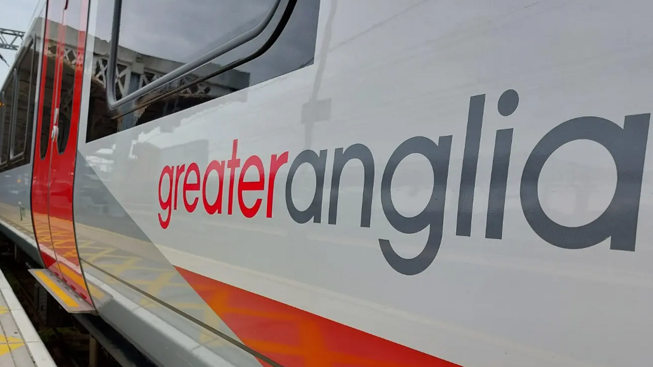A silver Greater Anglia train that has the company's logo on the side in red and grey. The train is stopped at a platform.