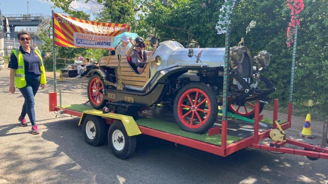 An old car, very similar to the one used in the film, is on the back of a trailer. Two children sit in the back and one waves for the camera.