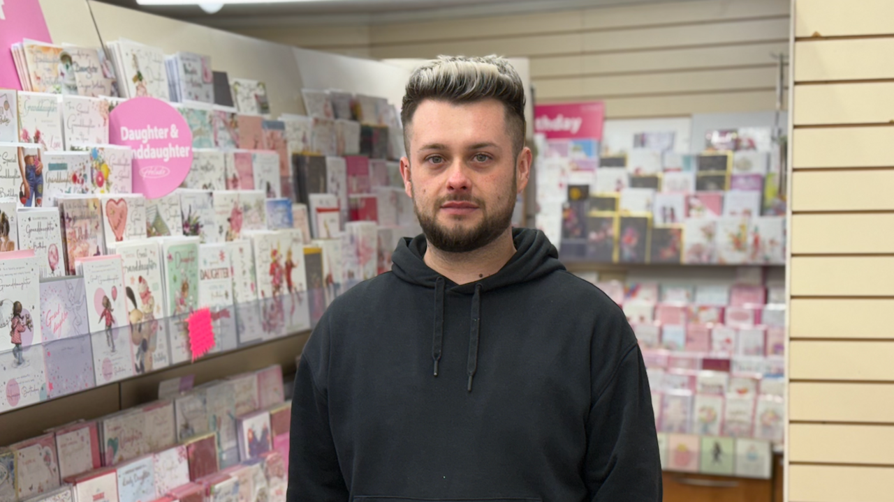 Callum McCall has short black and white hair. He is wearing a black hoody and is standing inside a card shop with greetings cards on display in the background.