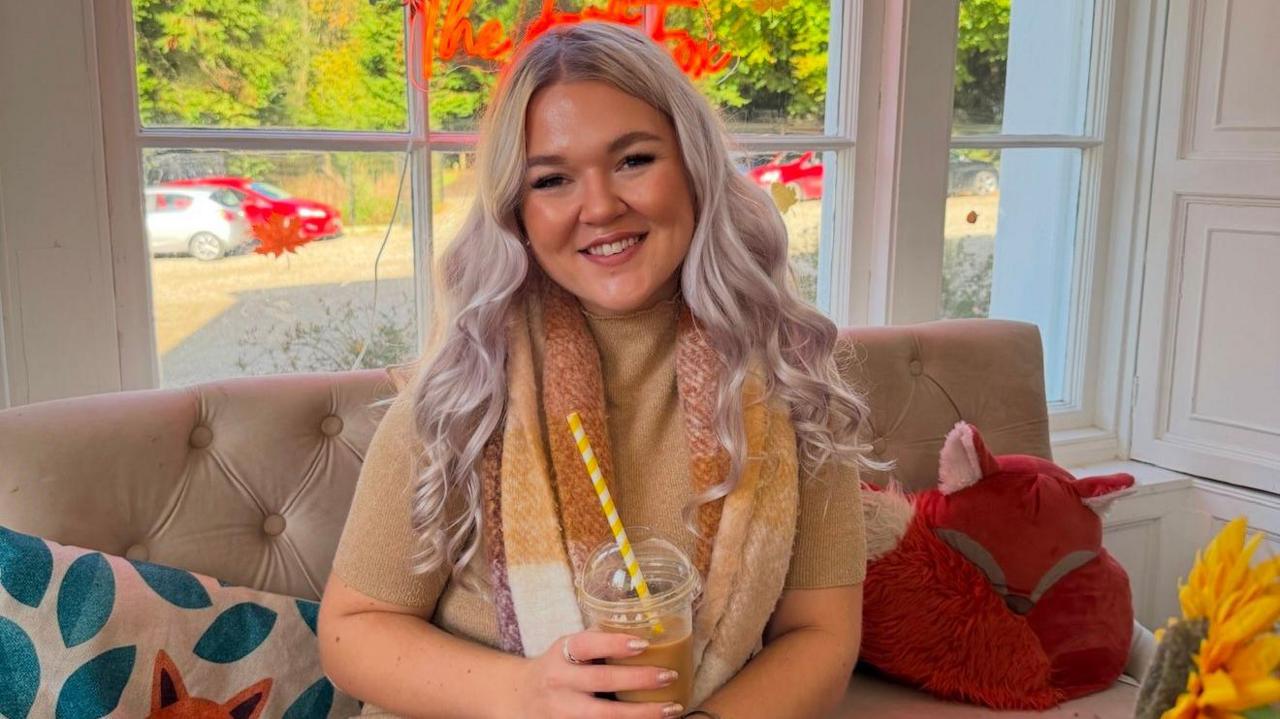 A woman sitting on a sofa in front of a window smiling at the camera. She is surrounded by colourful pillows. She is wearing a brown short sleeved jumper and a brown, yellow and white scarf. She is holding a plastic cup with a straw sticking out of it.