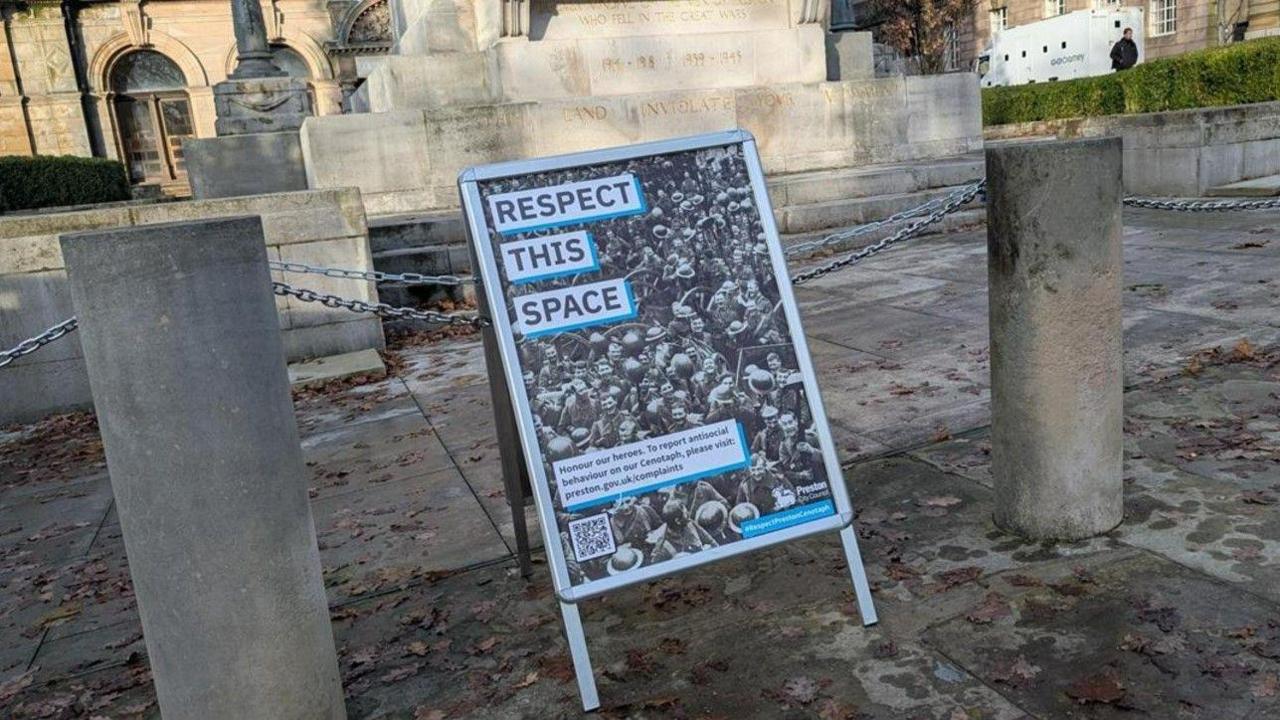 An A-board says 'Respect this space' and 'Honour our heroes. To report anti-social behaviour on our Cenotaph, please visit preston.gov.uk/complaints". There is a black and white image of soldiers on the A-board, with a Preston City Council logo and the hashtag #RespectPrestonCenotaph. The A-board is placed in front of the stone columns and chains that surround the Cenotaph in Preston. 