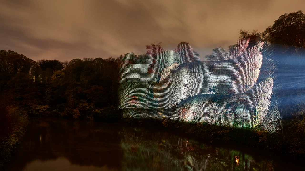 Geometrical Traces is an installation shining on the trees at Durham's riverside in narrow lines of green, yellow and purple, creating the impression of 3D-like wavy structure. The River Wear below slightly reflects the artwork's colours and the trees.