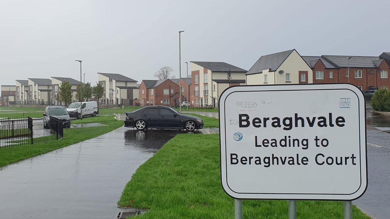 Shows a sign saying "Beraghvale leading to Beraghvale Court" in front of a grass verge, three parked vehicles and houses in the background.