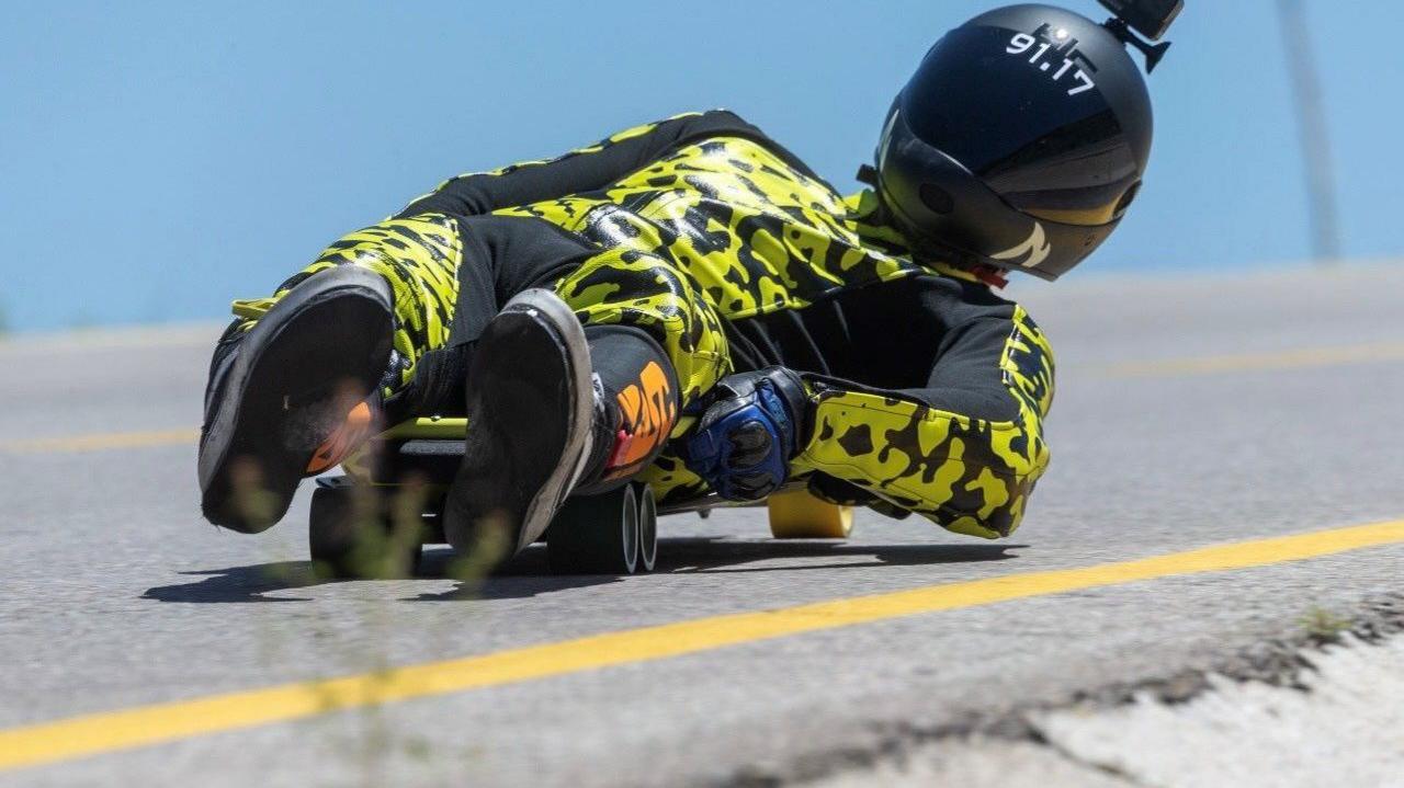 Man races down a hill on a skateboard. He is lying on the board, with his left elbow very close to the ground.
