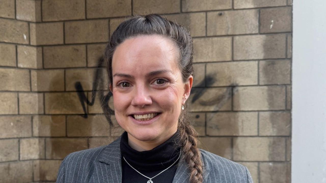 Photo of Fran Inman, chief executive at Cheltenham BID. She is smiling at the camera and wearing a dark grey pinstripe jacket and black top and behind her is a brown brick wall with one of the black spray-painted tags on it