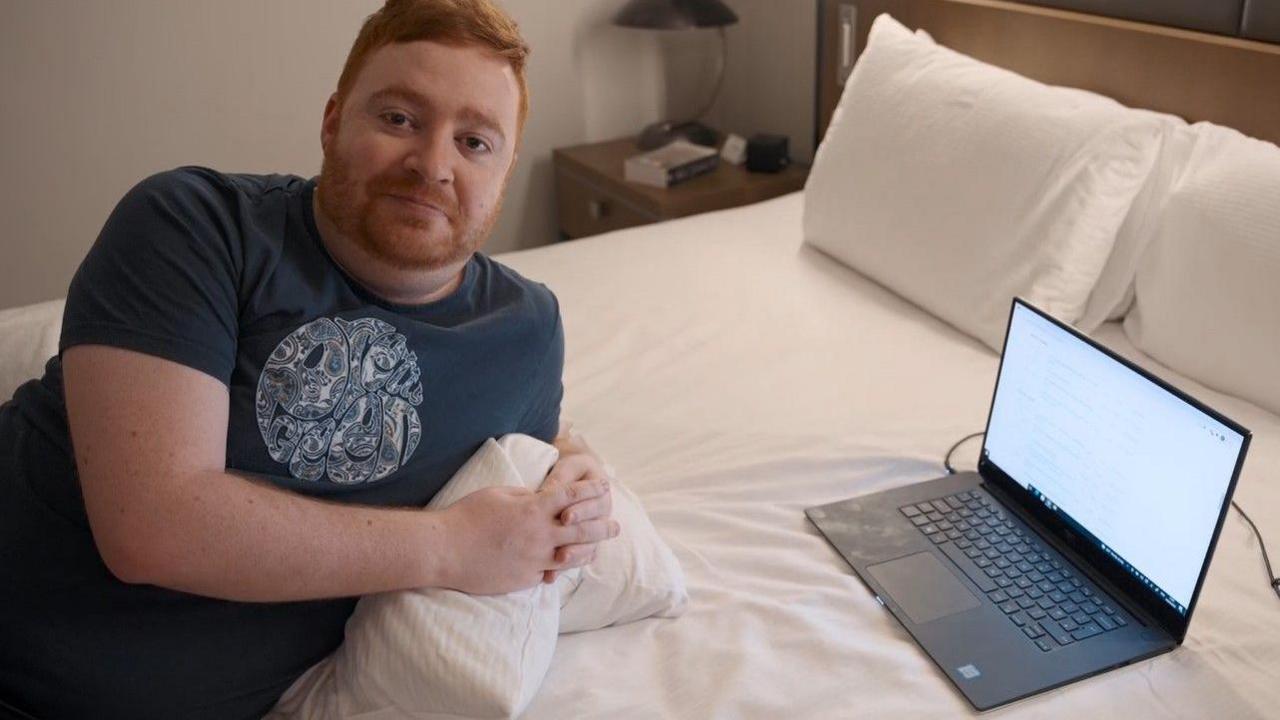 A man with ginger hair and a beard lies on a bed in front of a laptop computer