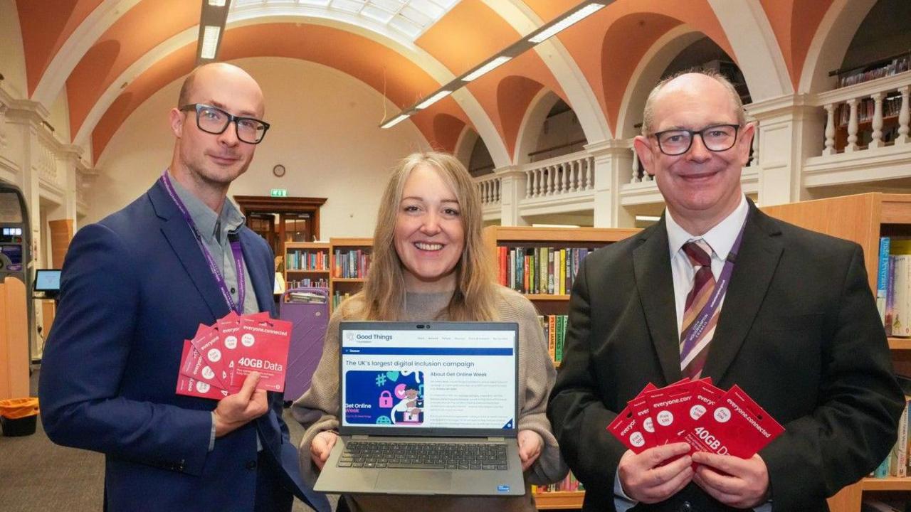 A man in a blue jacket and wearing glasses is on the left. A woman in the middle is holding a laptop. A man on the right with glasses is wearing a tie, white shirt and black jacket.