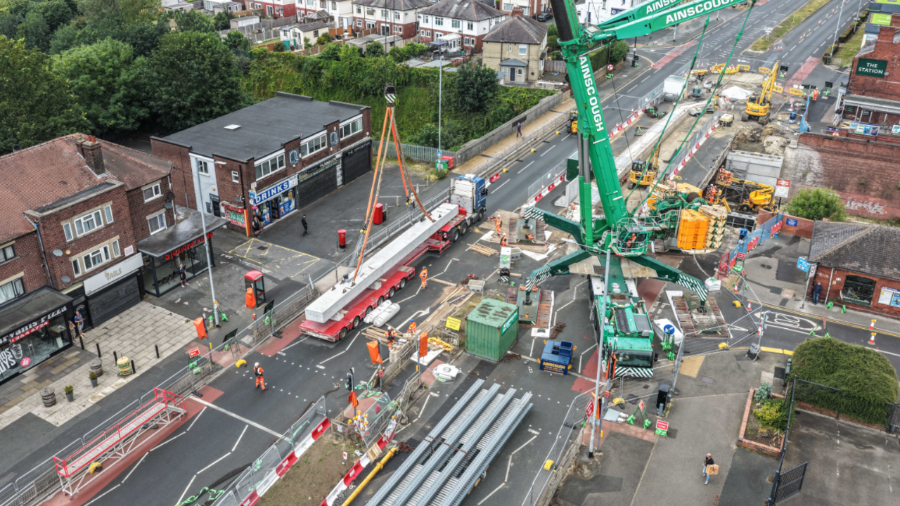 A birds eye view of a busy road blocked off by bollards and heavy machinery. A green crane towers over the scene. Shops line the road.