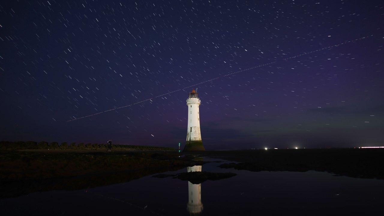 Dark blue night sky showing green and purple colours of the Aurora Borealis visible as a diagonal line of Perseid meteor passed over a white lighthouse 