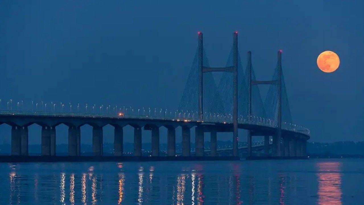 The Prince of Wales Bridge against a blue sky with a full orangey, red Moon to the right which is reflected, along with the lights from the bridge, in the estuary below. 