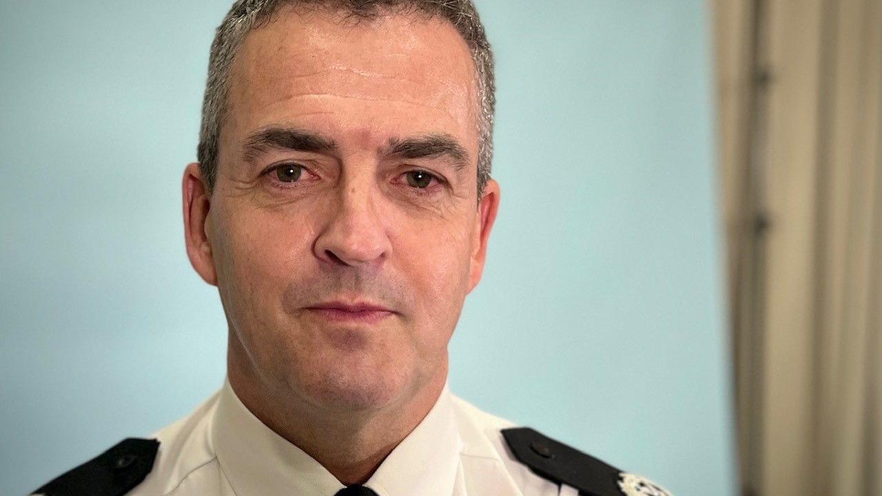 ACC Johnson in a head and shoulders shot, looking straight at the camera. He has very short grey hair and is clean-shaven. He is wearing a white police shirt with epaulettes.