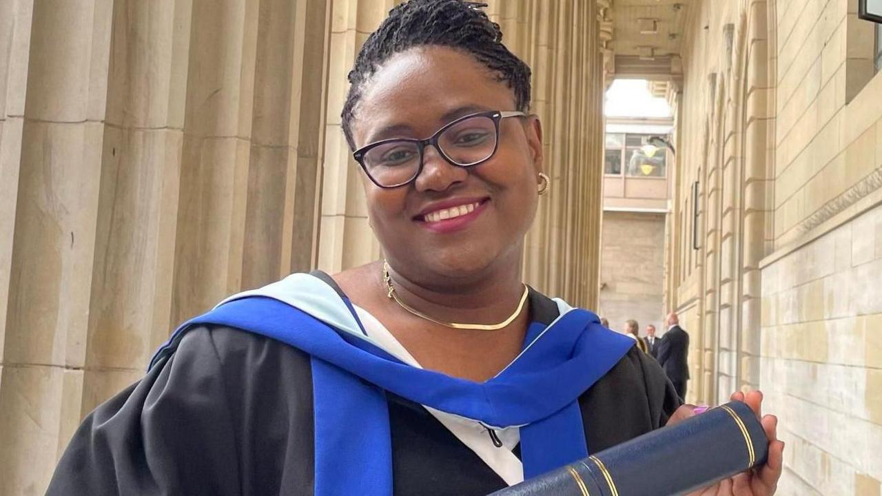 Dr Gomo, who is wearing black graduation robes, smiles as she holds a University of Dundee scroll. She is also wearing glasses.