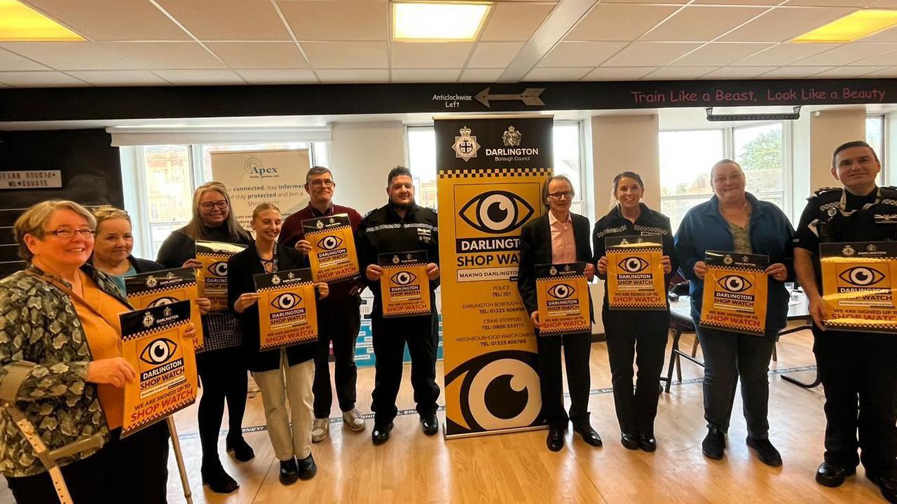 A group of 10 people standing in a line holding yellow signs. The signs have an eye on them with the words 'Darlington ShopWatch' underneath.