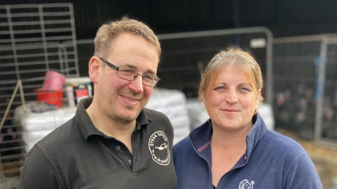 Richard and Tanza Dryden standing on their turkey farm. Richard has short brown hair and glasses. Tanza has brown hair  which has been tied up. She is wearing a blue fleece. a flock of turkeys stand in an enclosure behind them.