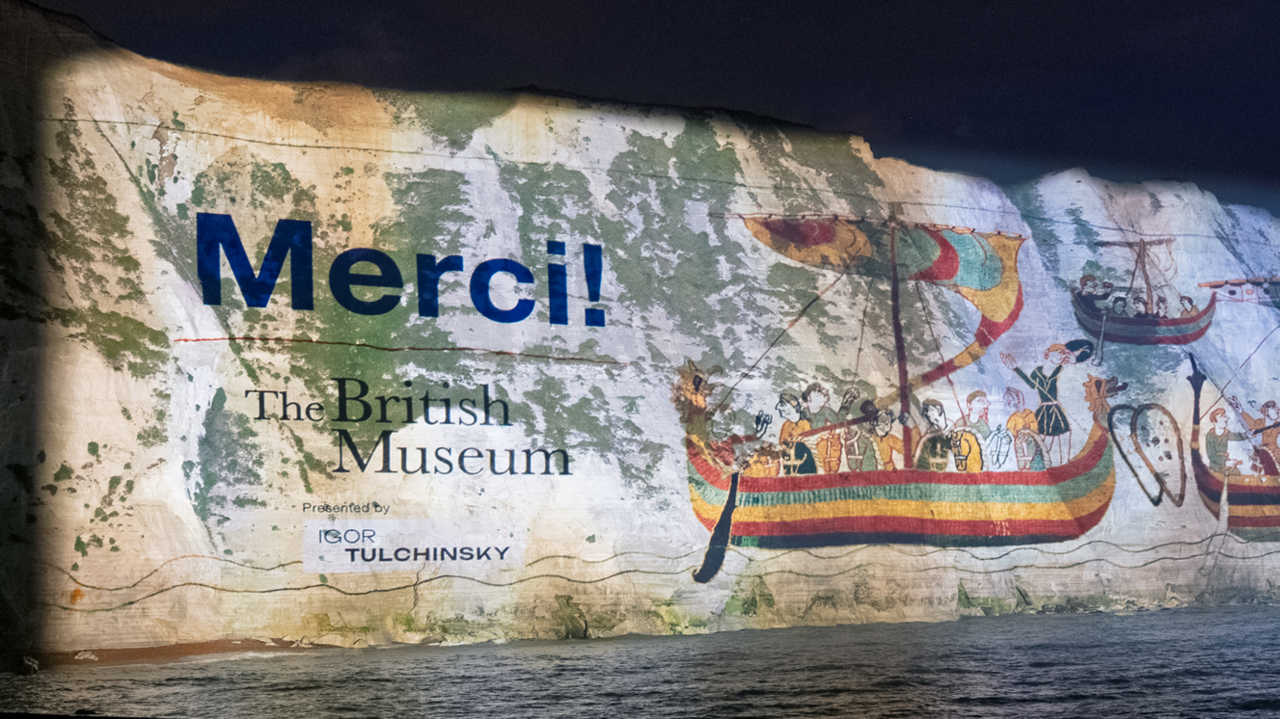 White cliffs of Dover at night, covered in a giant projection with images of wooden boats from the tapestry, and "Merci"