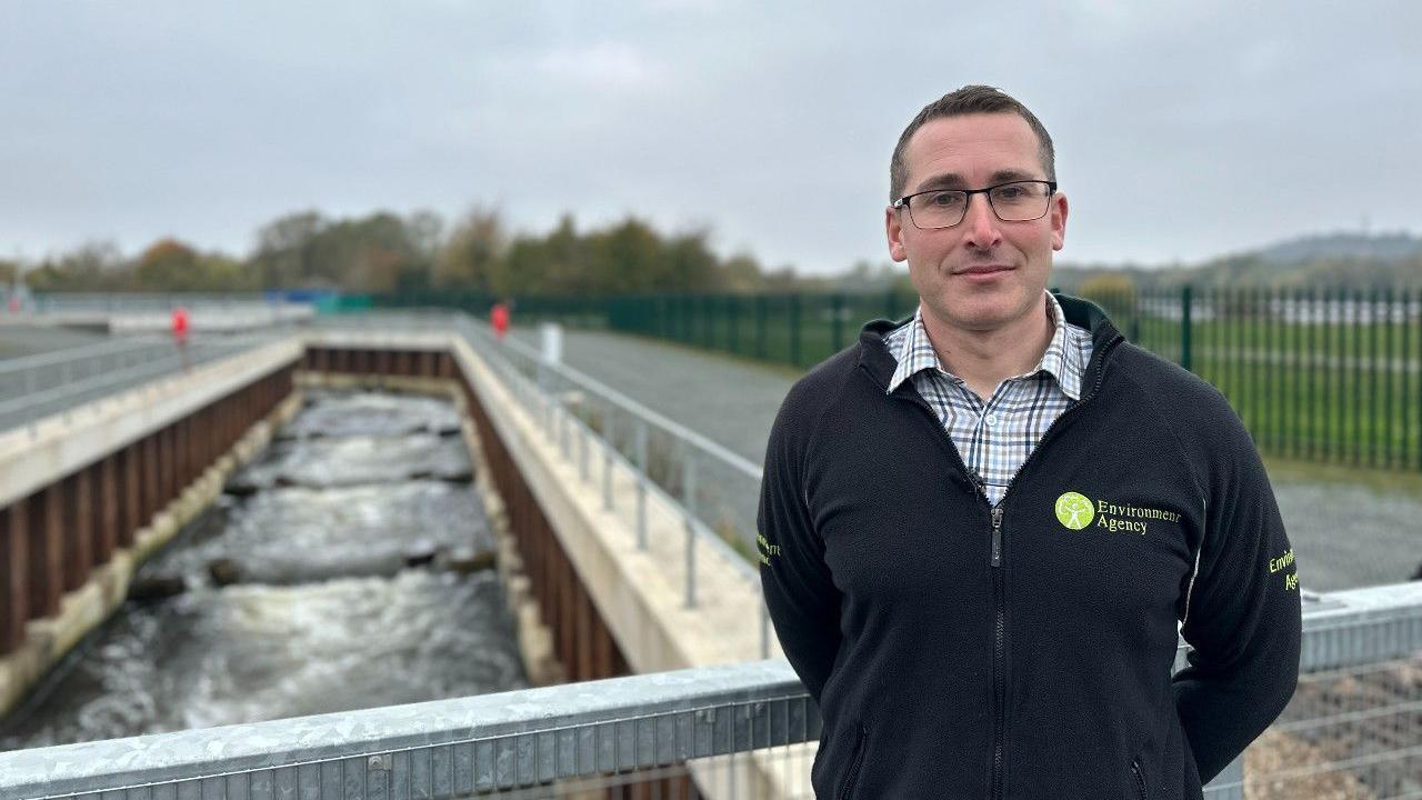 Steve Lawrie, wearing an Environment Agency fleece, stood on a bridge, with the fish pass channel flowing beneath