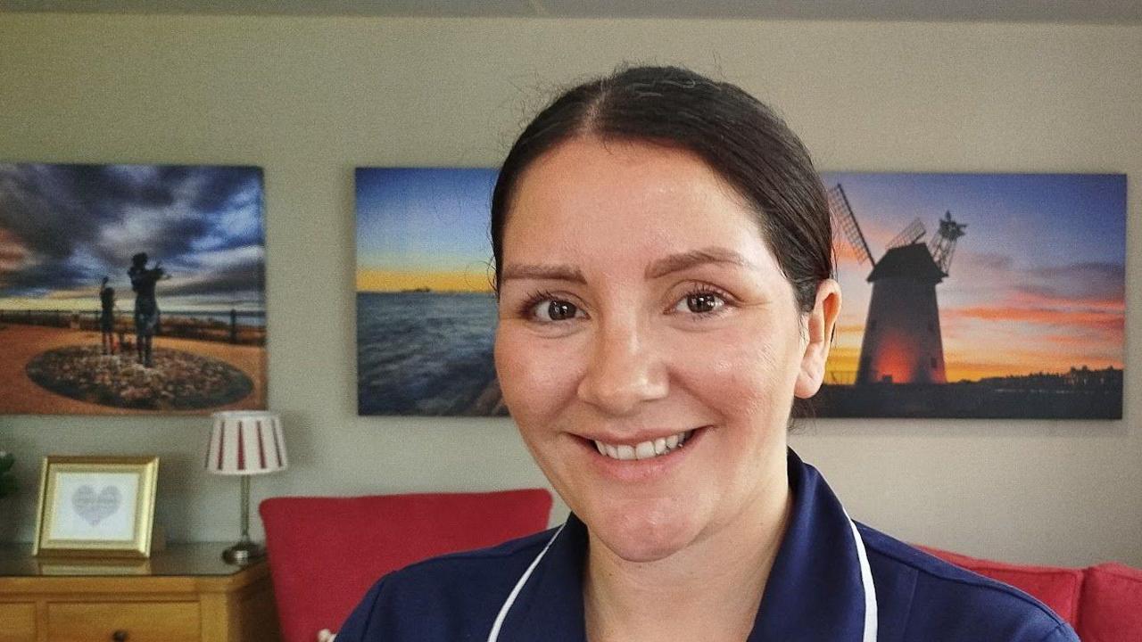 Megan Parry-Jones has dark hair tied back in a bun and is wearing a blue nurse's uniform. In the background, behind her, photos of the Fylde Coast hang on a grey wall.