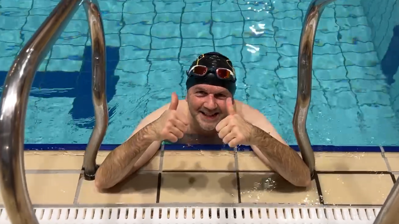 Michael Coombes, a man wearing a swimming cap and leaning on the edge of an indoor pool, gives a thumbs up.