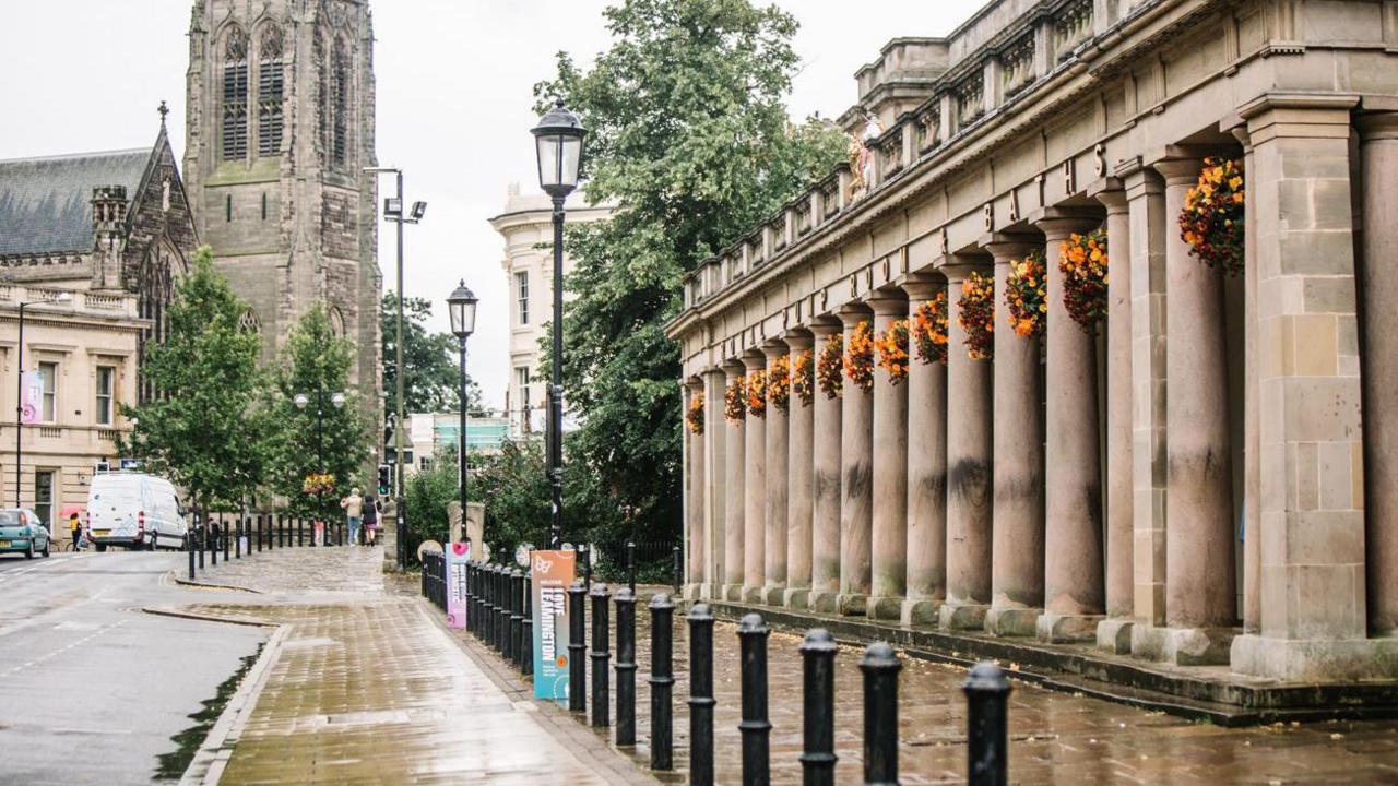 A grey columned building on the right side of a street with a row of black bollards in front of it and a church in the distance