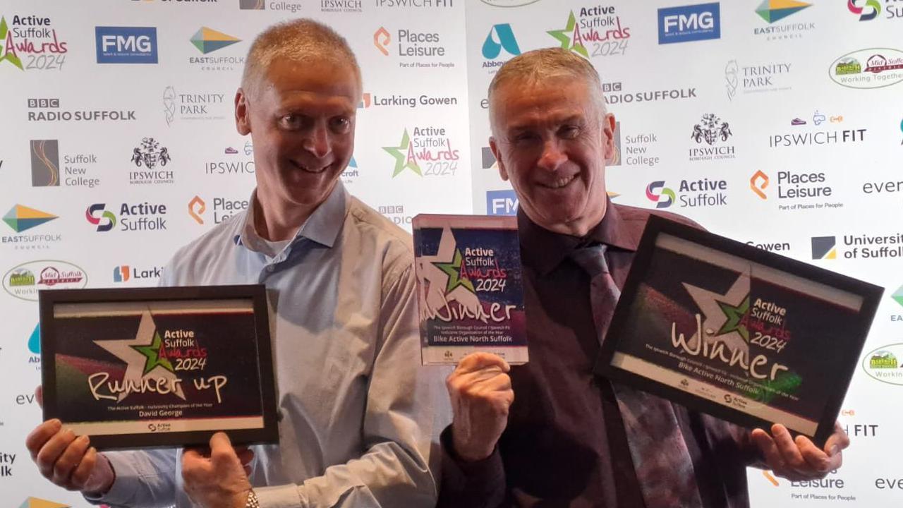 David George smiles at the camera holding a certificate. He has short grey hair and wears a blue shirt. Standing next to him is Phil Robbins who holds two other certificates and smiles at the camera. He also has short grey hair and wears a purple shirt with a purple tie.