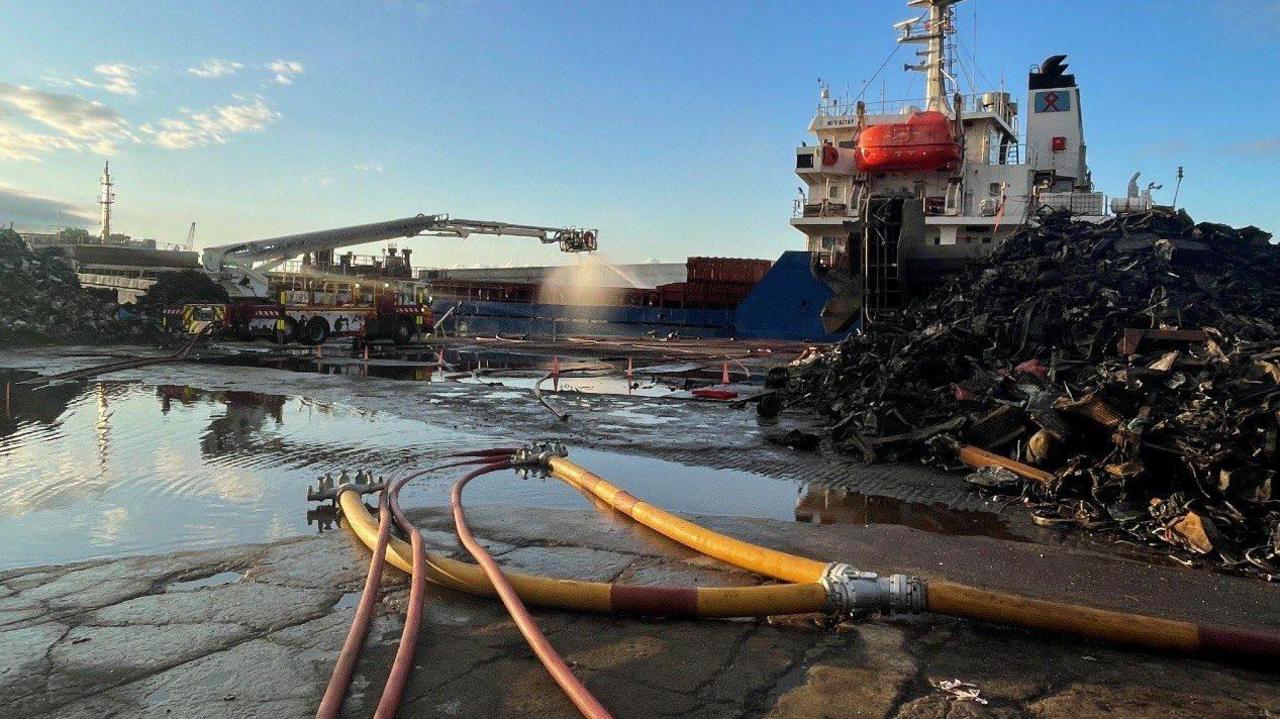A long-armed aerial platform sprays water over the scene of the fire, with hoses in the foreground and a blue and white ship in the background