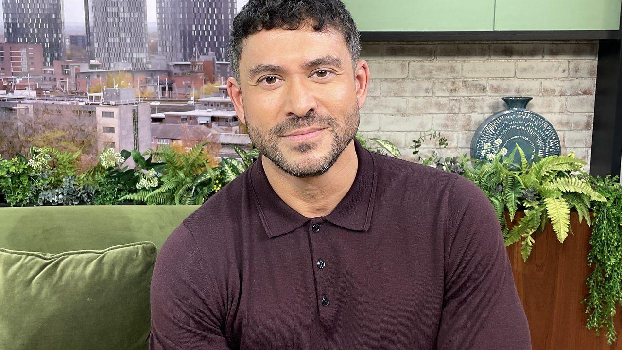 Rav Wilding is a male with close-cropped dark hair and facial hair. He wears a dark burgundy polo shirt with long-sleeves and sits on the green velvet Morning Live studio sofa. 