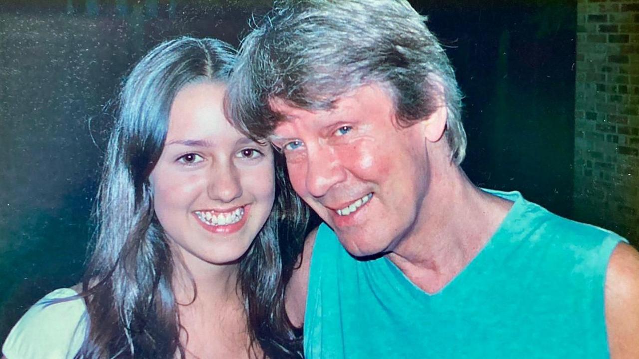 Teenage Laura has long brown hair and is pictured smiling with her father who has his arm round her. He is wearing a blue sleeveless shirt and is smiling. He has short greying hair.