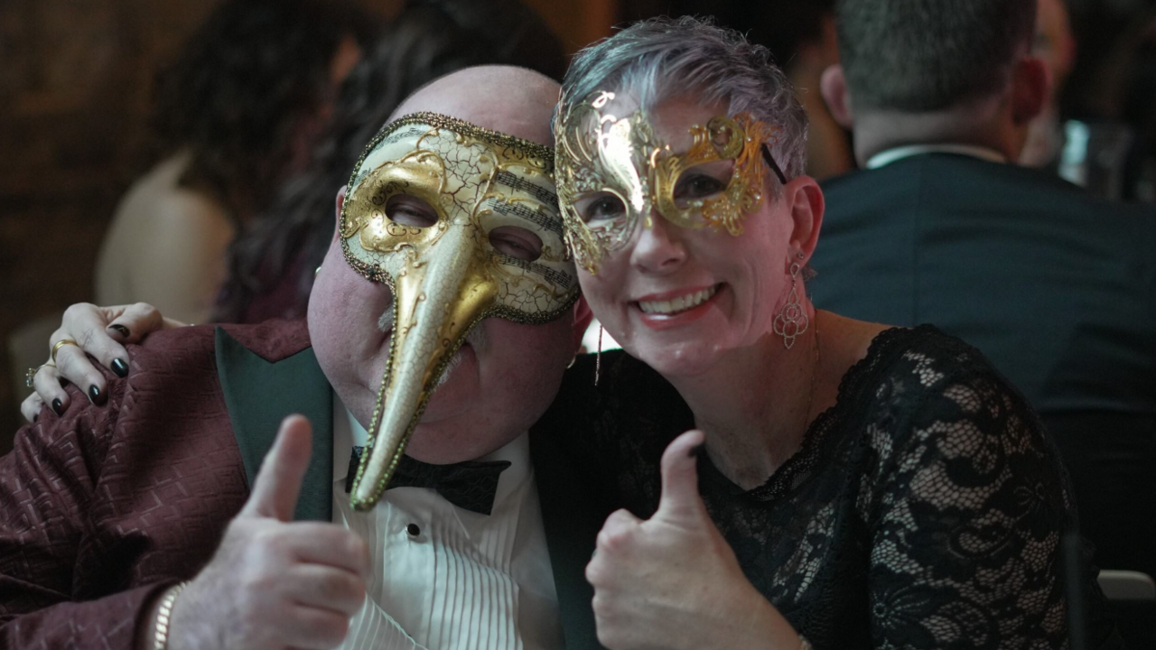 A close-up of an older man and a woman at a table, both with gold masks on and smiling to the camera with their thumbs up. The man is wearing a smart burgundy tuxedo with green lapels and a white shirt and green bow tie. The lady is wearing a black lace top