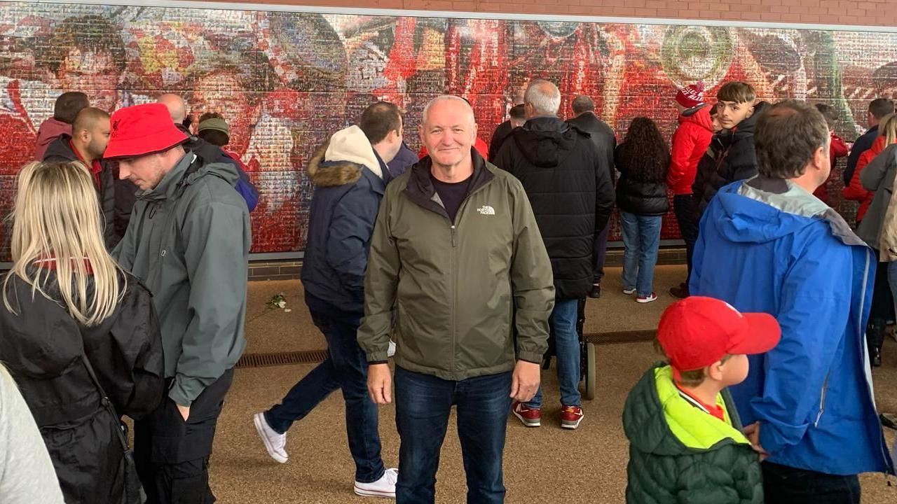 A bald man, wearing a green coat and jeans, stands in the middle of the frame while other people walk or stand around him. Behind him on the wall is a mural of past Liverpool FC players.