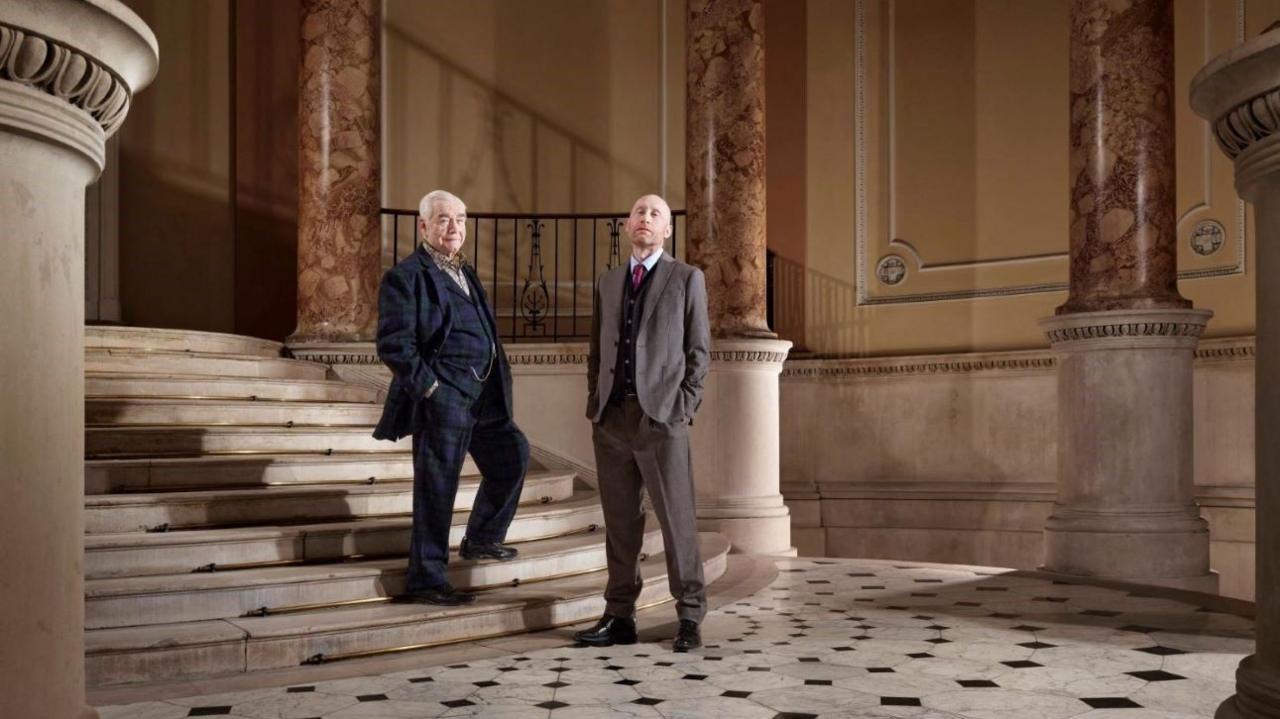 Brian Cox and Sandy Grierson pose in suits in a Georgian room full of marble columns.