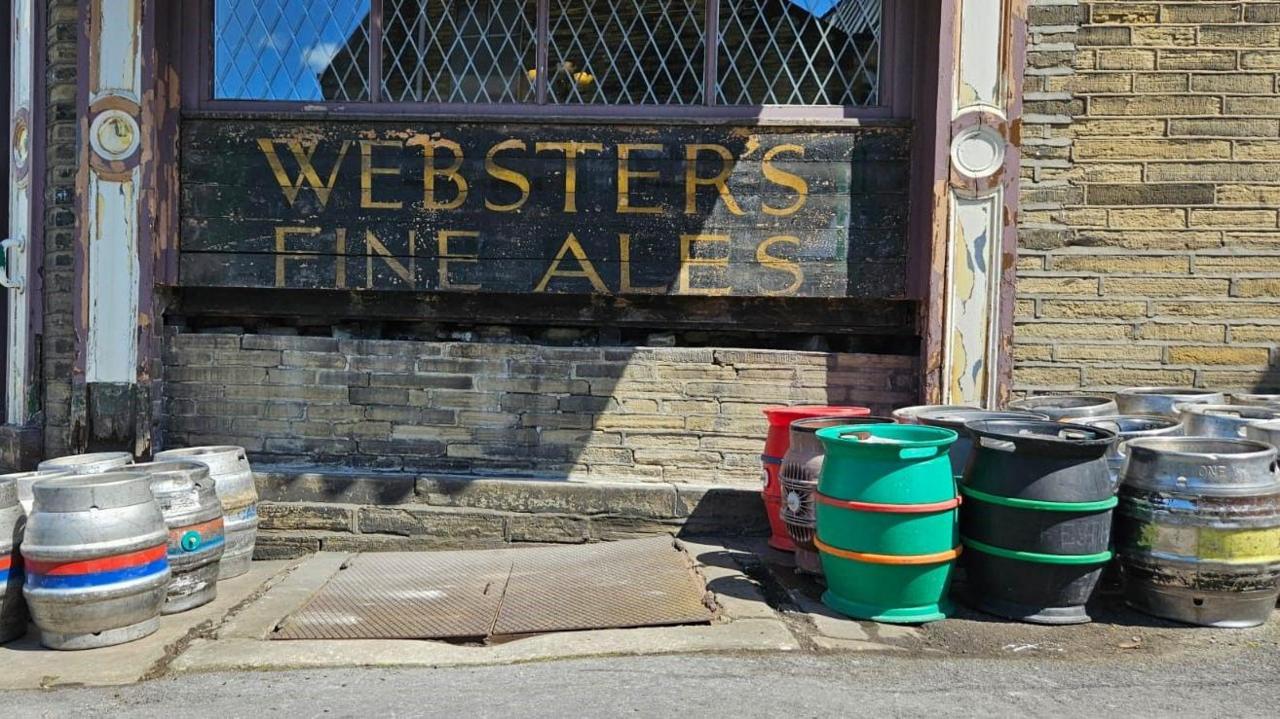 A pub cellar hatch with metal doors over it with beer metals either side on the pavement and a fade brown painted sign on wooden boards with the words Webster's Fine Ales spelled out in gold lettering. 