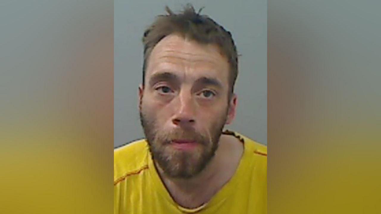 Mugshot of a man with a brown beard and scruffy hair, wearing a yellow T-shirt