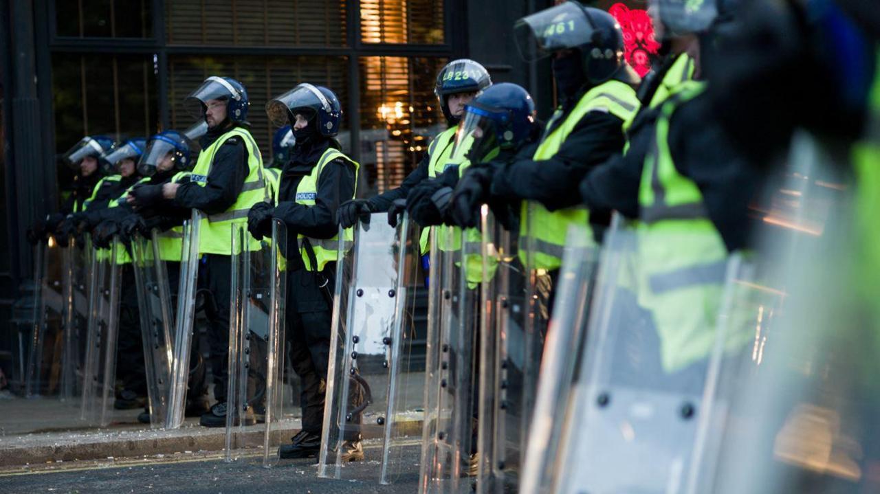 A line of police officers wearing riot gear. They are holding clear shields in front of them.