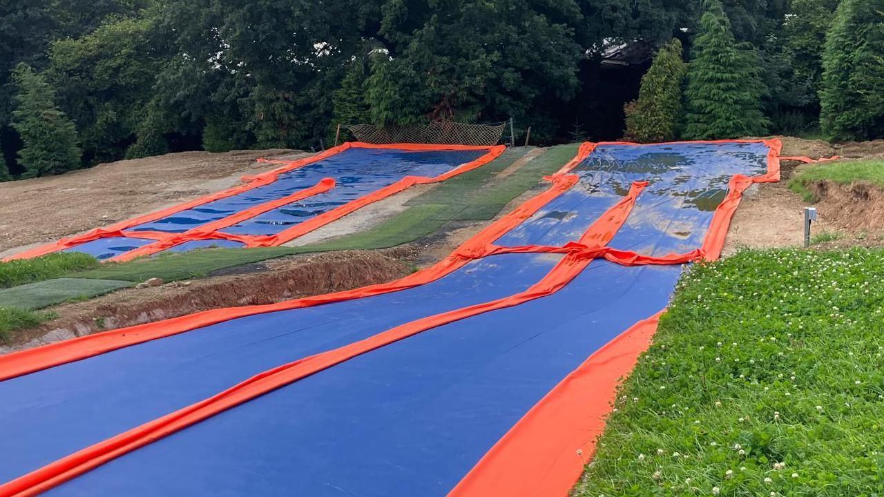 Two blue and red slip and slides laid out on a green field with green fir trees behind them.