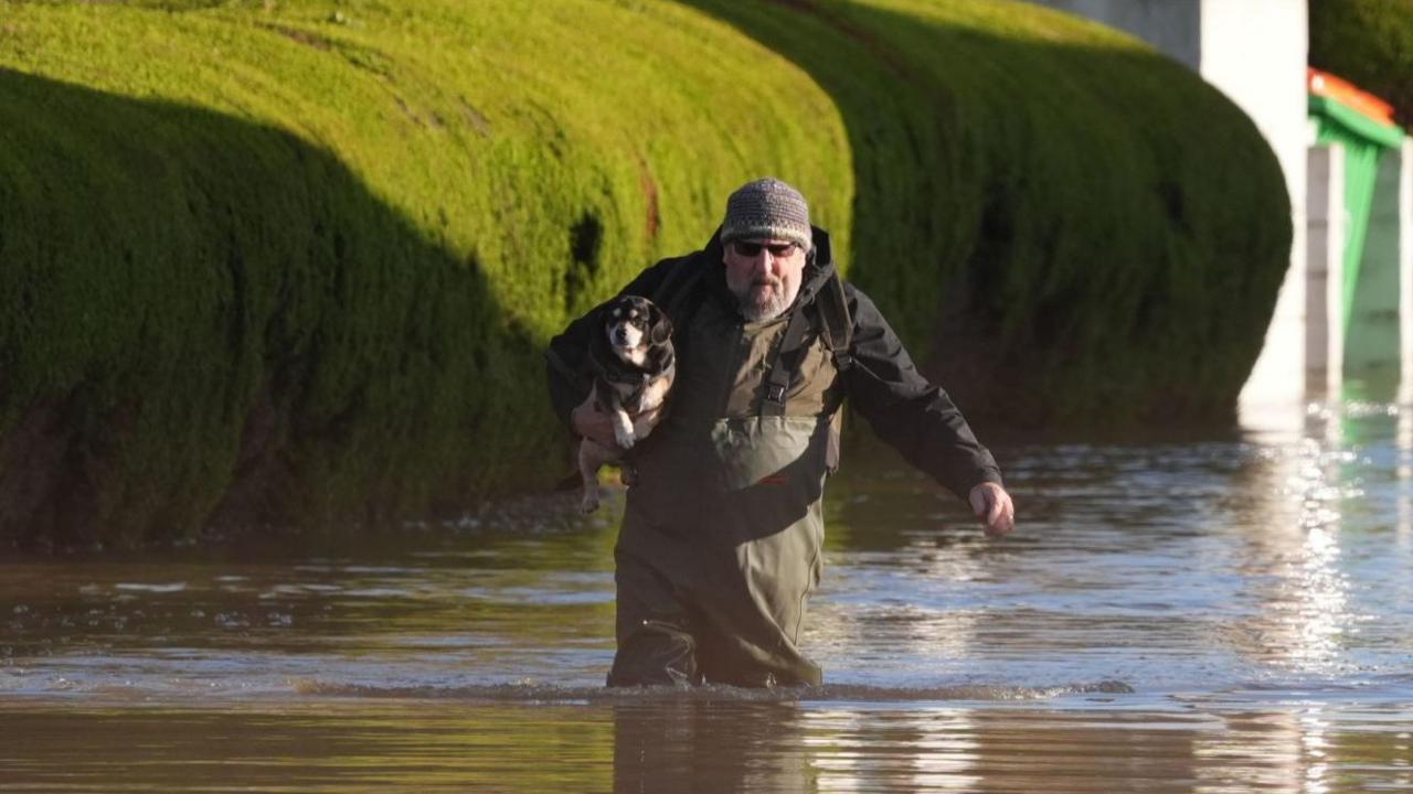 A man wearing waders carries a dog at The Little Venice caravan park in Yalding, Kent, which is surrounded by rising flood water