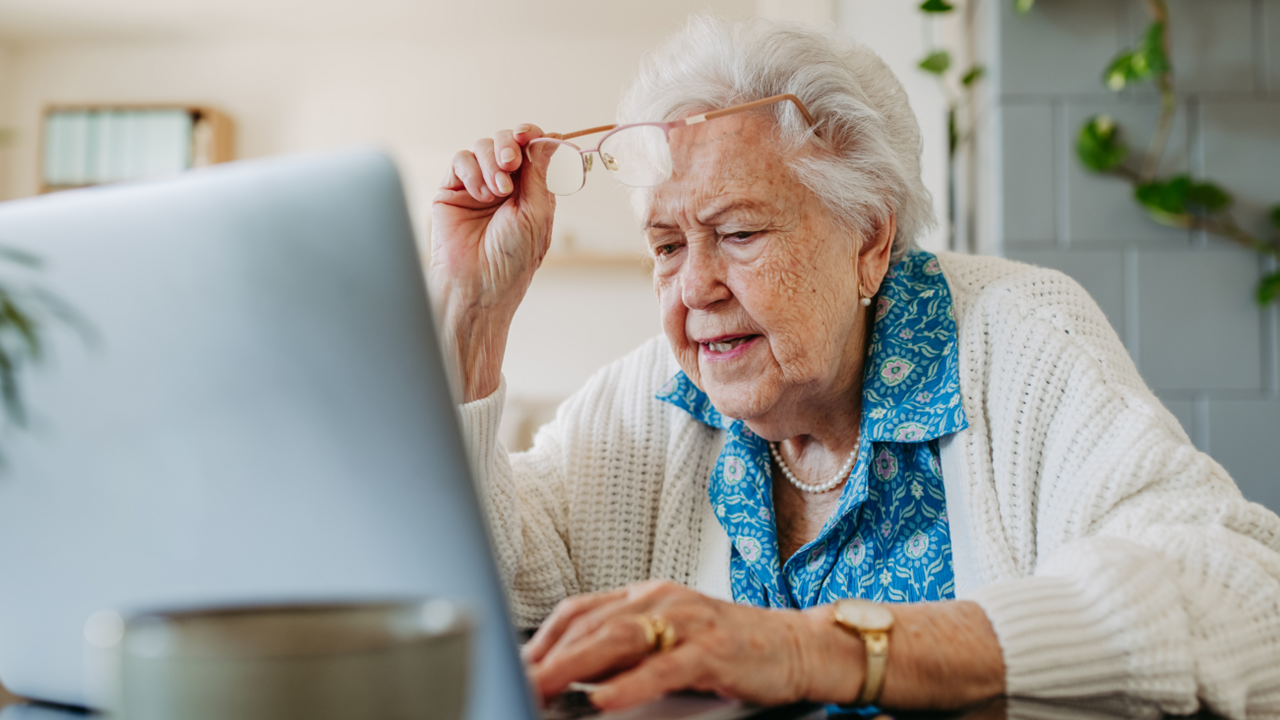 A woman with grey hair, blue blouse and white cardigan lifting up her glasses and looking at a computer monitor