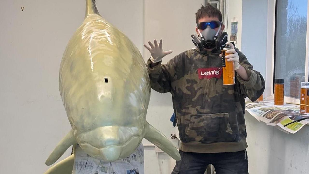 A person in a brown camouflage hoodie wearing a respirator facemask posing for the camera next to the dolphin which has its nose facing towards the camera. The person is holding up a spray can and has their other hand held up. The dolphin is covered in pale cream coloured primer.