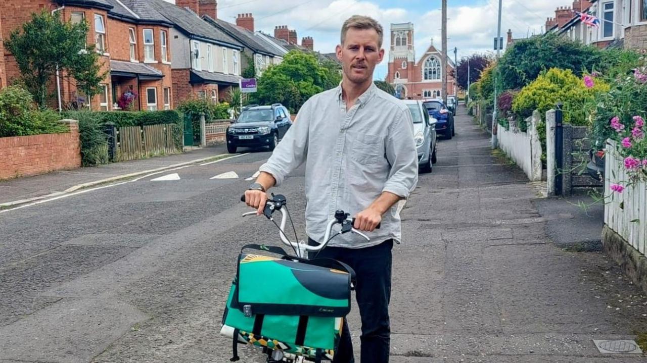 Andrew McClean standing next to his bike on a residential street. He has light brown hair, wearing a light blue shirt and black trousers and watch.