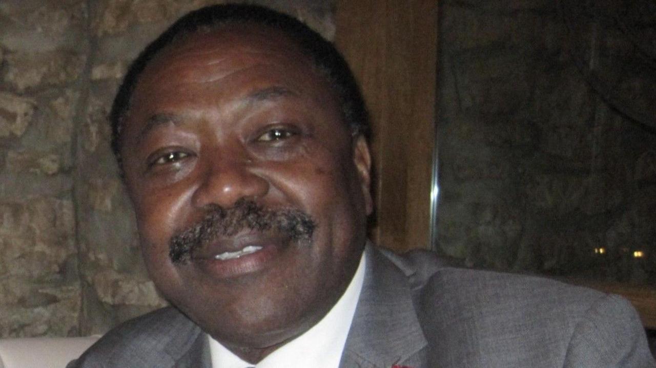Professor Christopher Balogun-Lynch, looking at the camera and smiling. He has a moustache, and is wearing a white shirt, with a grey jacket. He is sitting in front of a stone wall. 