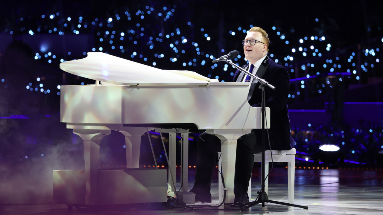 A man in a suit singing on stage while sat behind a large white piano