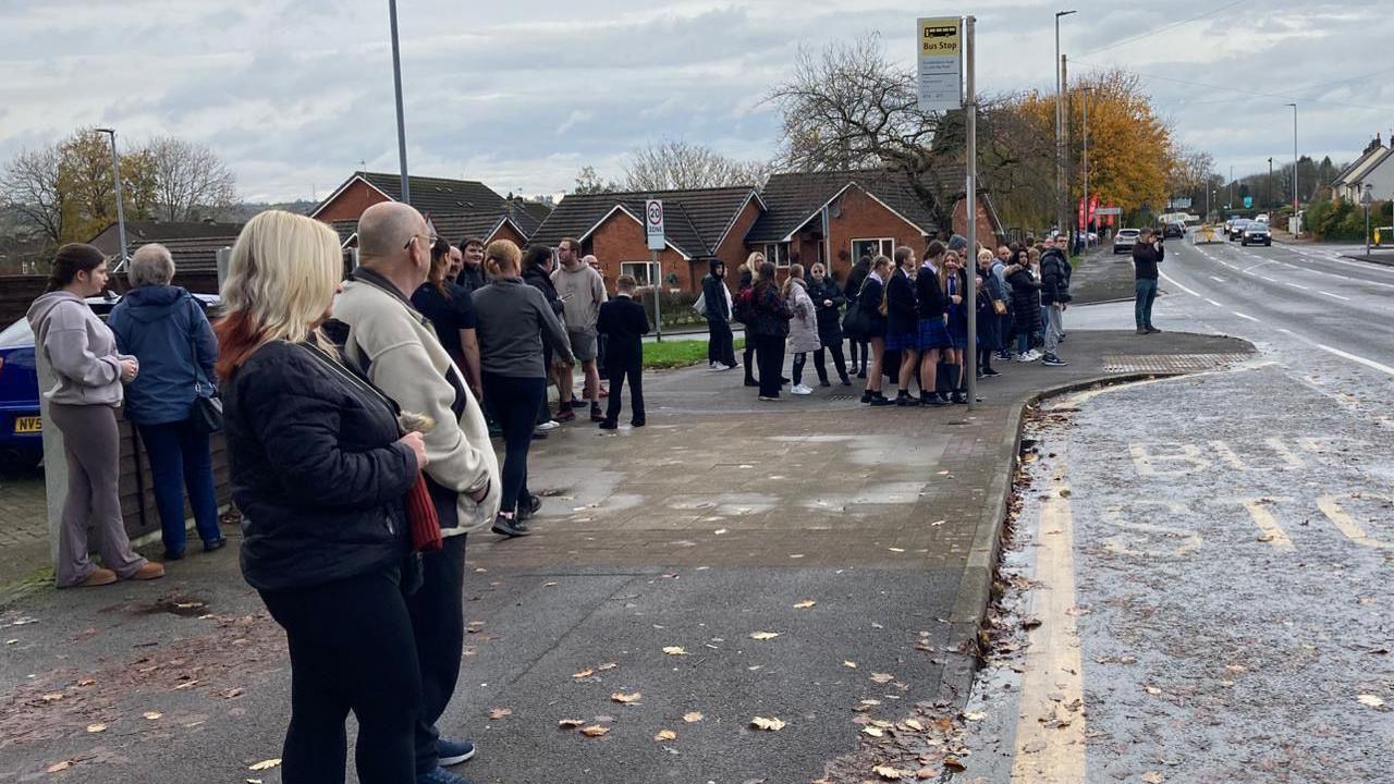 A crowd of people are pictured at the crossroads where Keith once worked, waiting for the procession to arrive. There is a bus station lay-by in the photo and a woman and man stand front of frame whilst a crowd of school children stand further back.