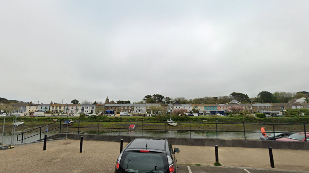 South Quay in Hayle on a grey day. The water runs below a car park. There is a row of houses on the opposite bank. A car is parked in the foreground behind bollards.