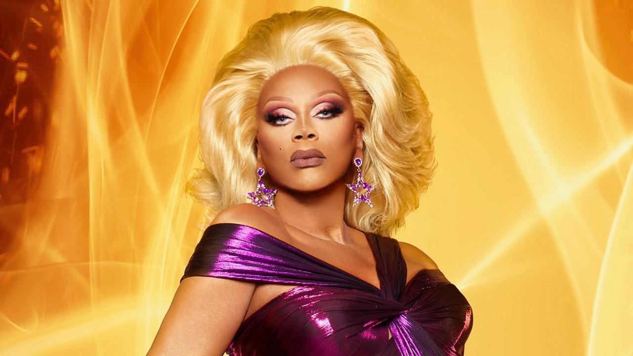 RuPaul wearing a blonde wig and a purple dress stood in front of a orange or yellow background