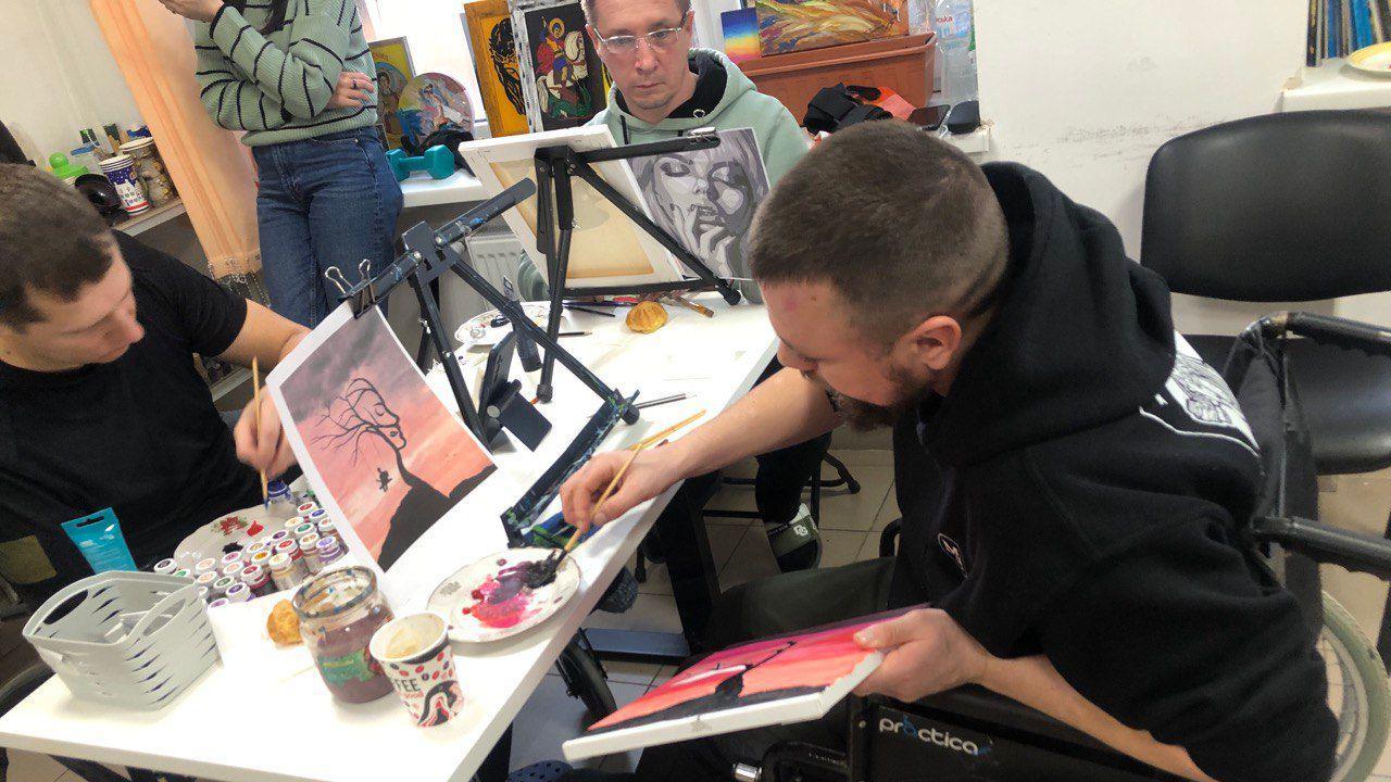 Three men paint at a table. The man closest is in a wheelchair.