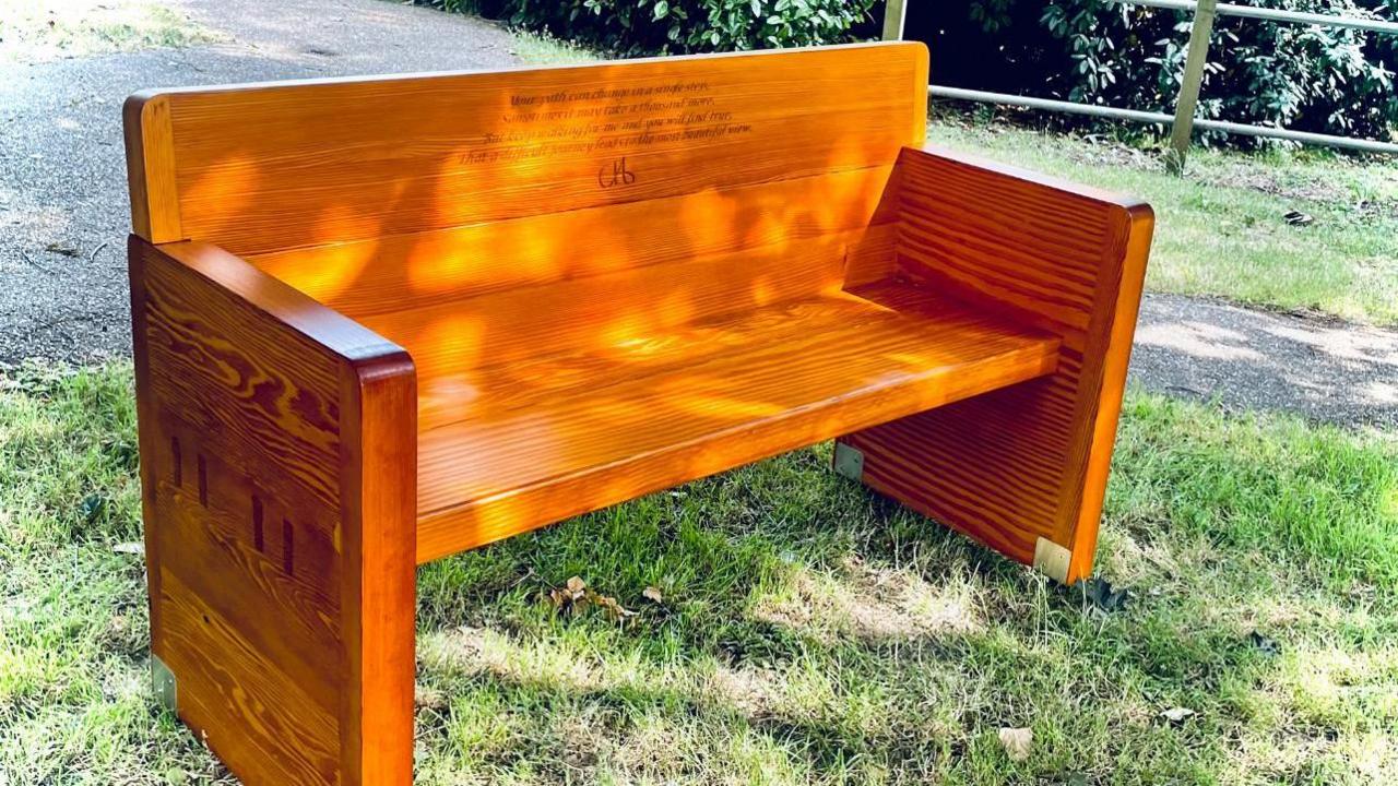 A light-brown bench stands on grass. It has a high back containing an inscription.